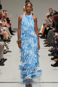 Model walks the runway in a floor-length blue-and-white fringe dress with gold accessories.