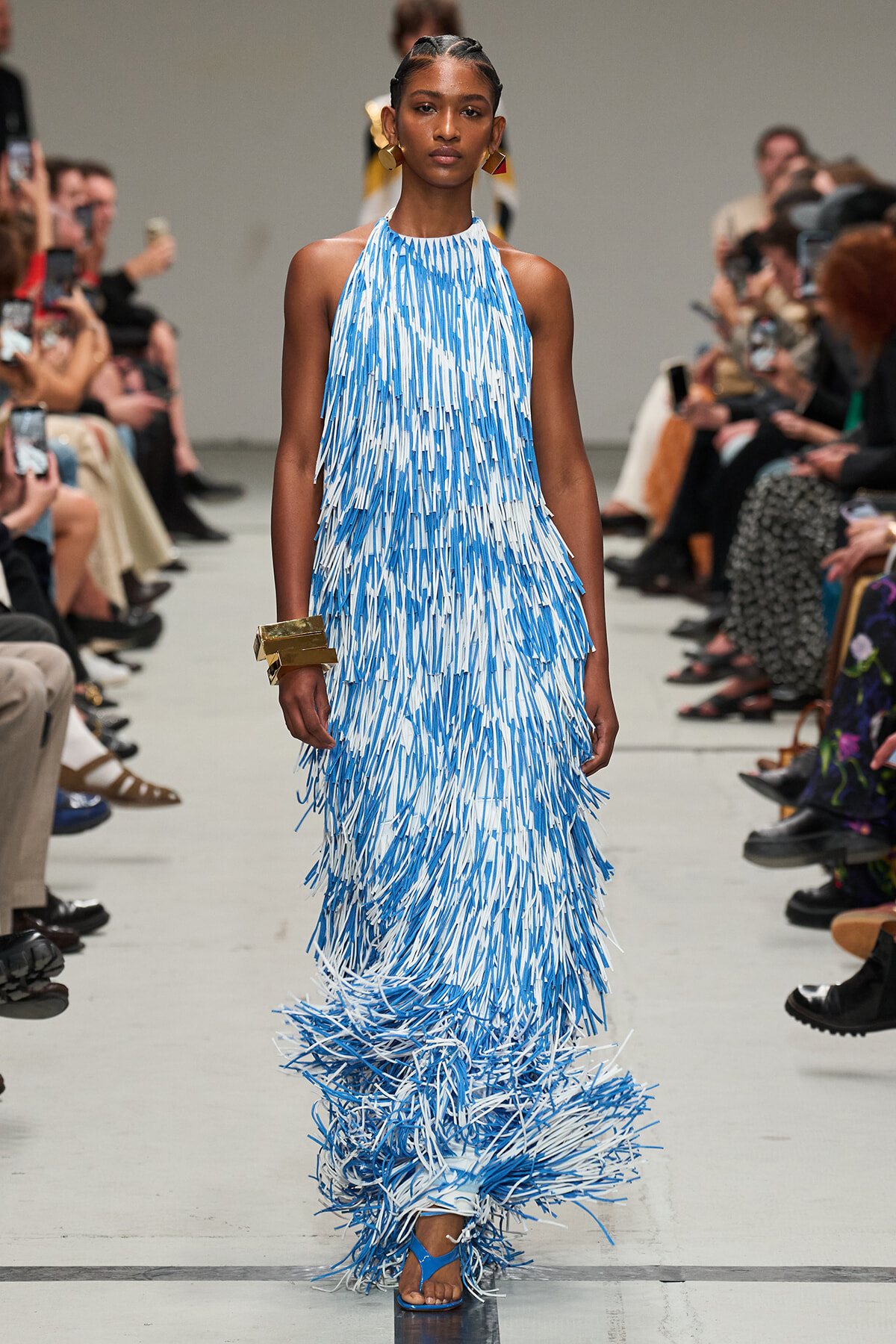 Model walks the runway in a floor-length blue-and-white fringe dress with gold accessories.