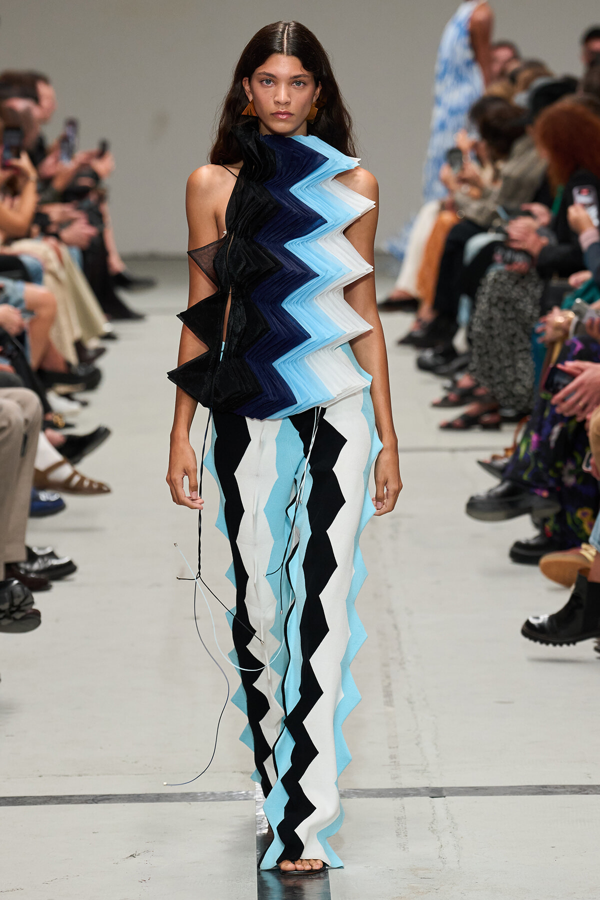 Model walking a fashion runway in a zigzag layered dress in shades of blue, black, and white.