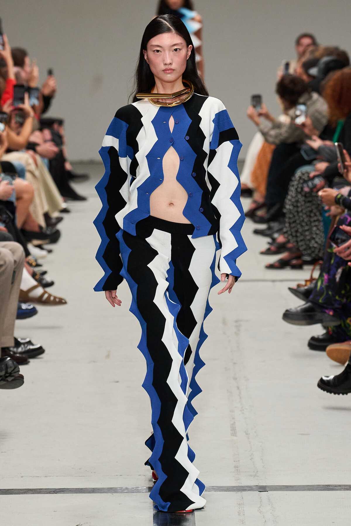 Model walks the runway in a blue, black, and white zigzag knit suit with an exposed midriff at a fashion show.