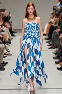 Model walking a runway in a strapless blue-and-white geometric dress with fringe-length panels and a chunky gold necklace.