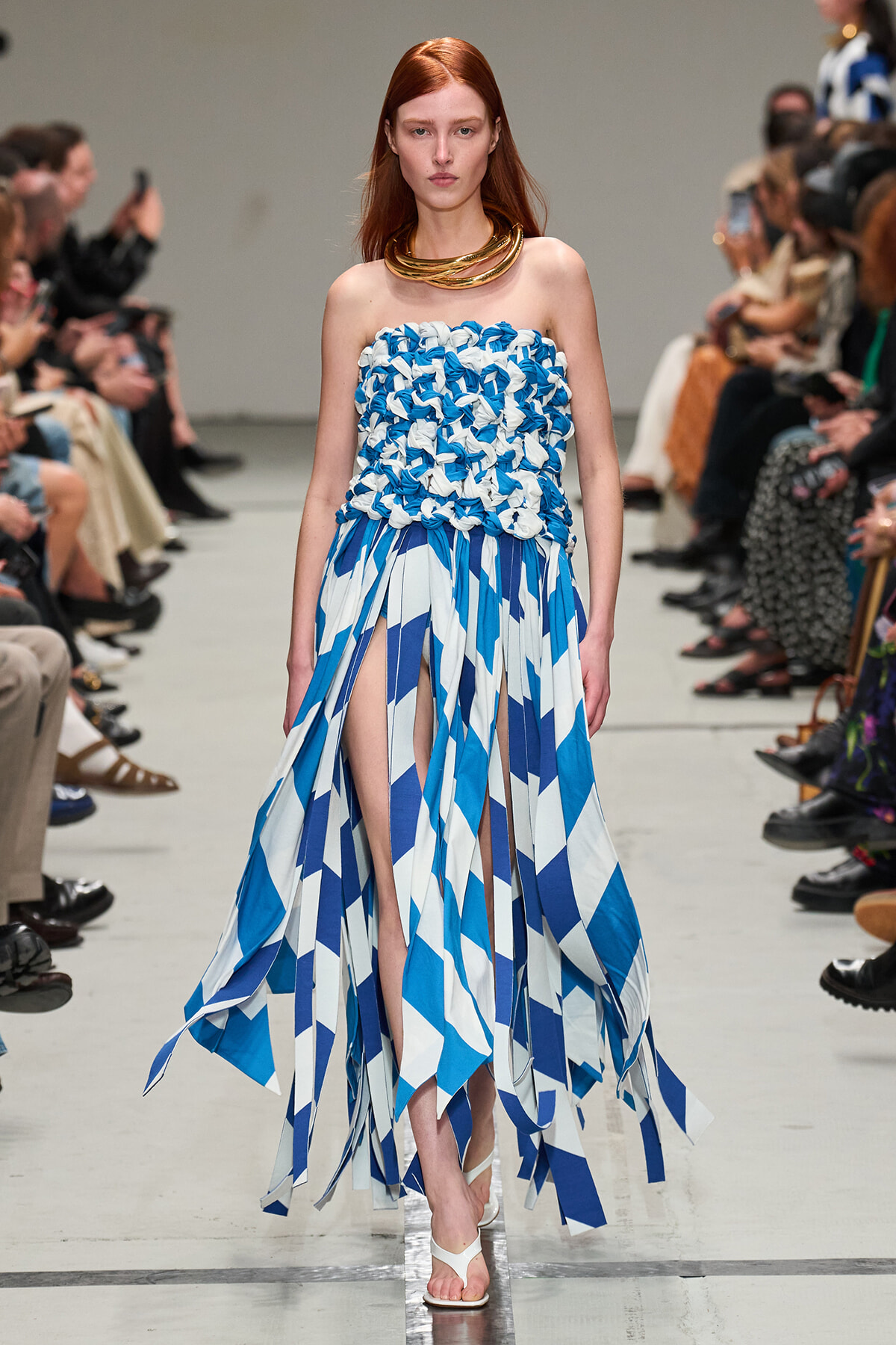 Model walking a runway in a strapless blue-and-white geometric dress with fringe-length panels and a chunky gold necklace.