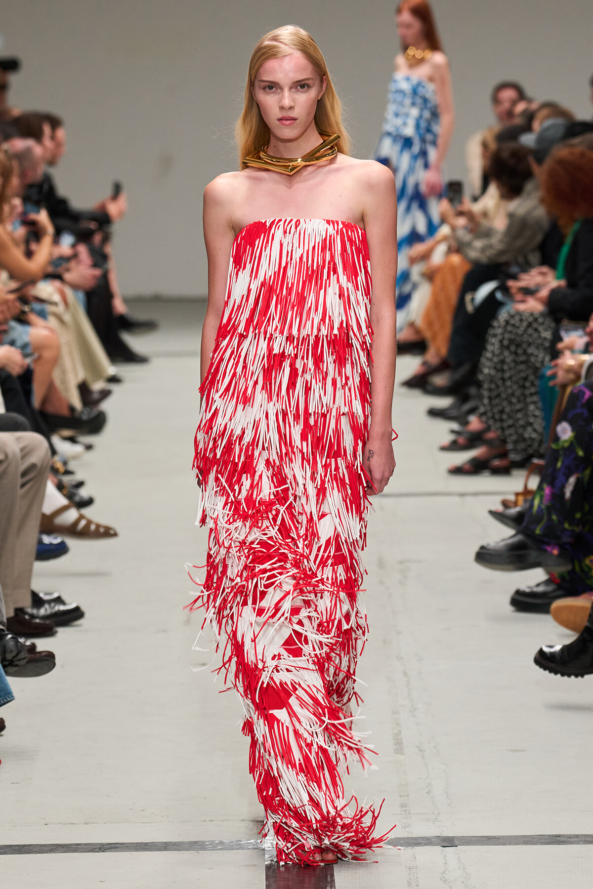 Model walks the runway in a strapless red-and-white fringe gown with a chunky gold neckpiece; audience watches from the sides.