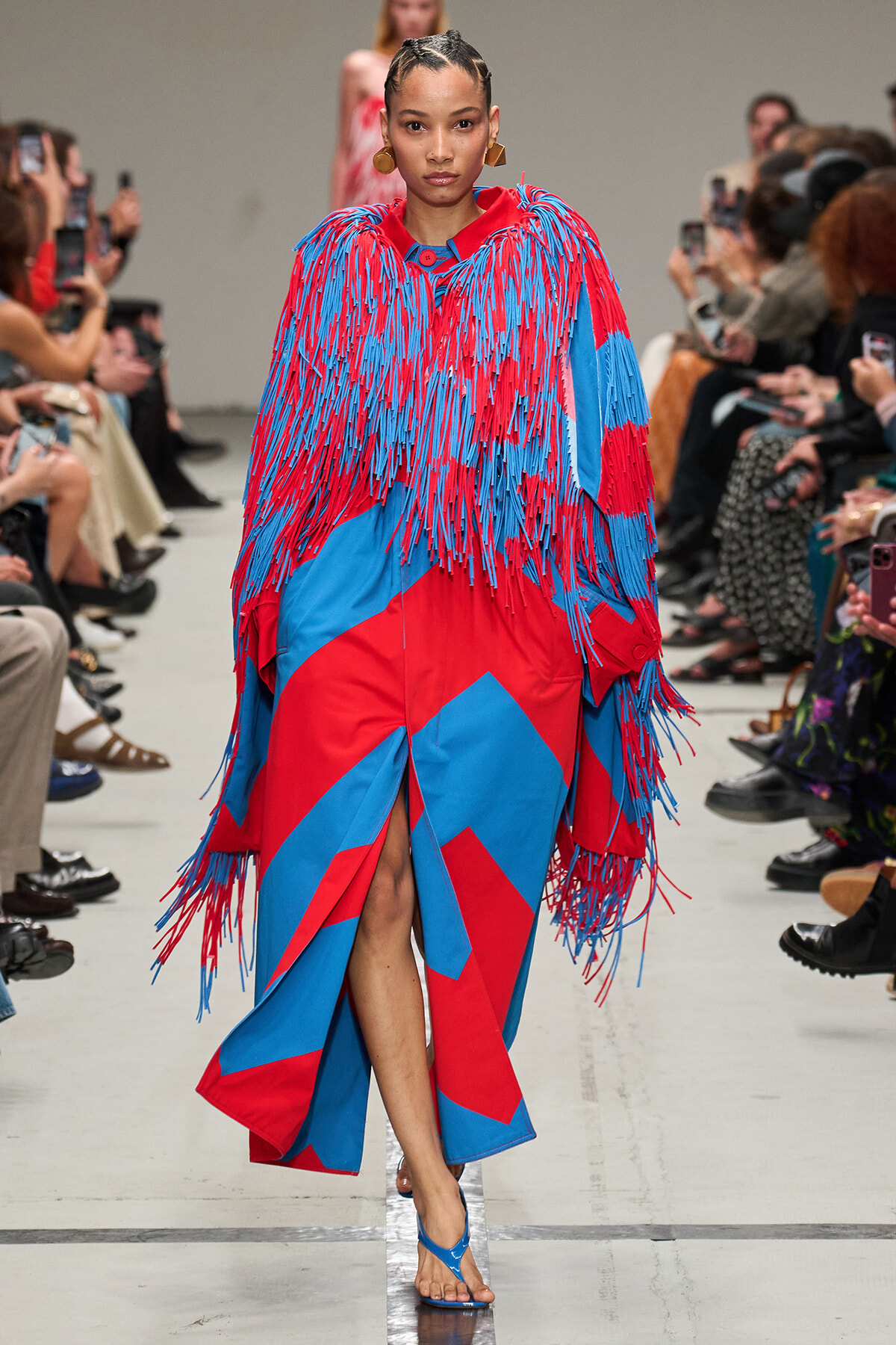 Model walks a runway in a red-and-blue fringed coat with geometric color blocks, paired with blue sandals; audience watches from sides.