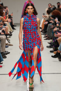 Model walks the runway in a blue and red woven dress with long dangling fabric strips, audience on both sides.