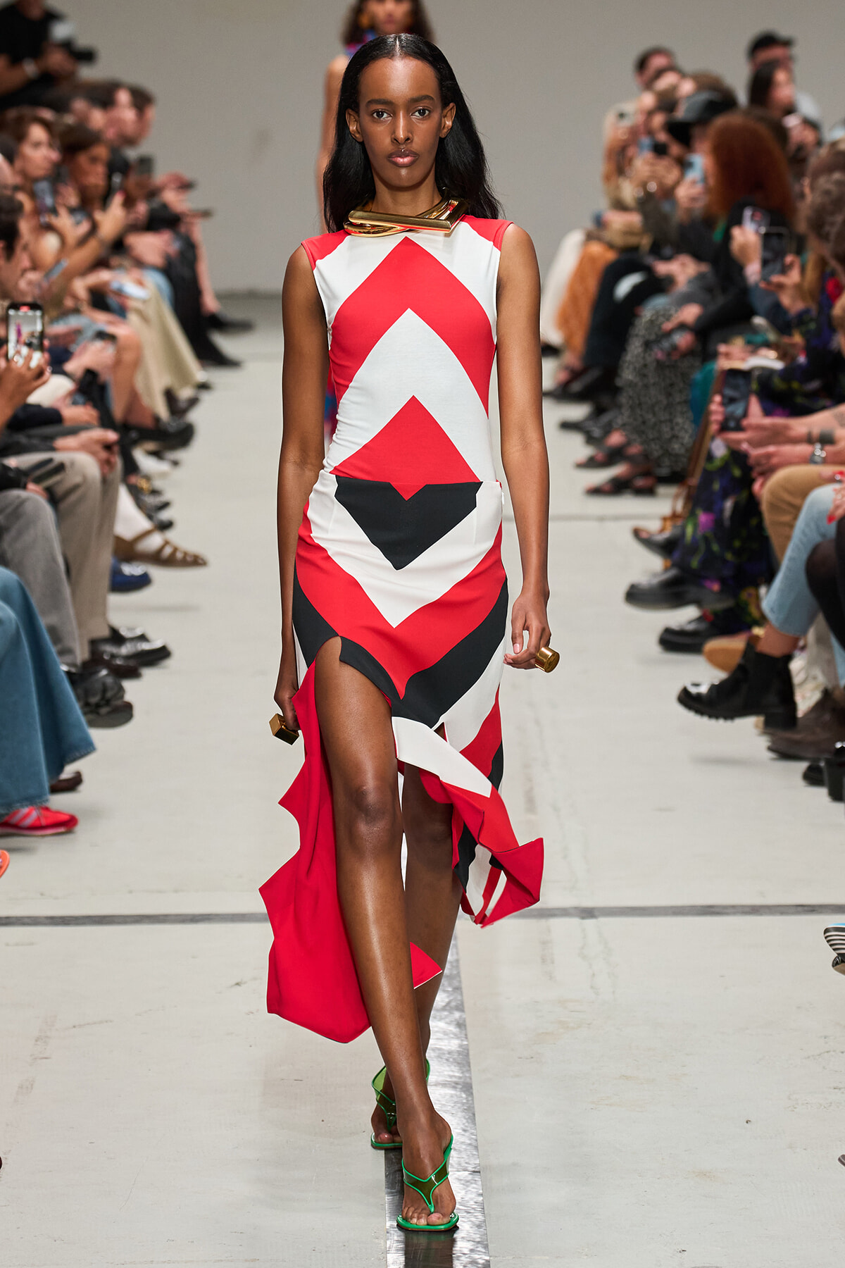 Model walks a runway in a bold red, white, and black geometric dress with an asymmetric hem; bright green strappy heels.