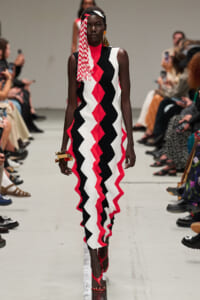 Model walking a fashion runway in a sleeveless red, black, and white zigzag dress with a matching headband and gold earrings. Audience seated along the catwalk.