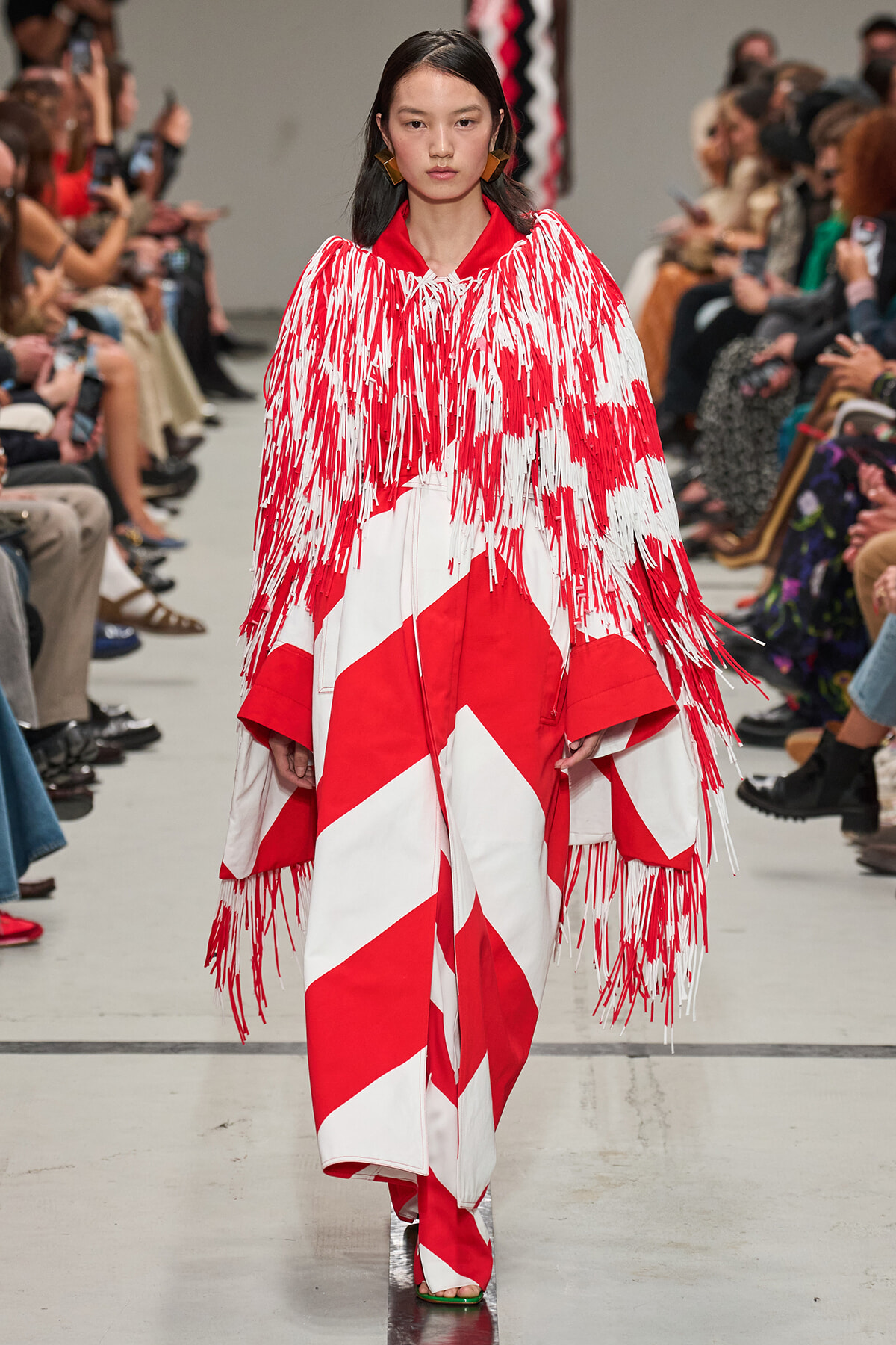 Model walks runway in a bold red-and-white fringe cape over a geometric red-and-white dress, with large gold earrings and red lips.