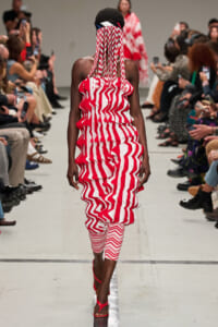 Model walks the runway wearing a red-and-white zigzag dress with long fringe over her face and a dark headpiece.