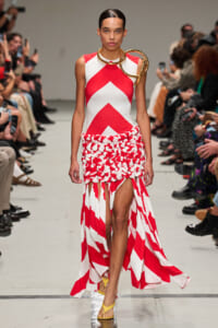 Model walks the runway in a red-and-white chevron dress with a textured, braided skirt and oversized gold circular neck jewelry; audience lines the runway in the background.
