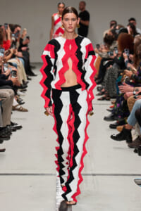 Model walking runway in a red, black, and white zigzag knit suit with an open midsection and matching pants; audience lines the runway behind.