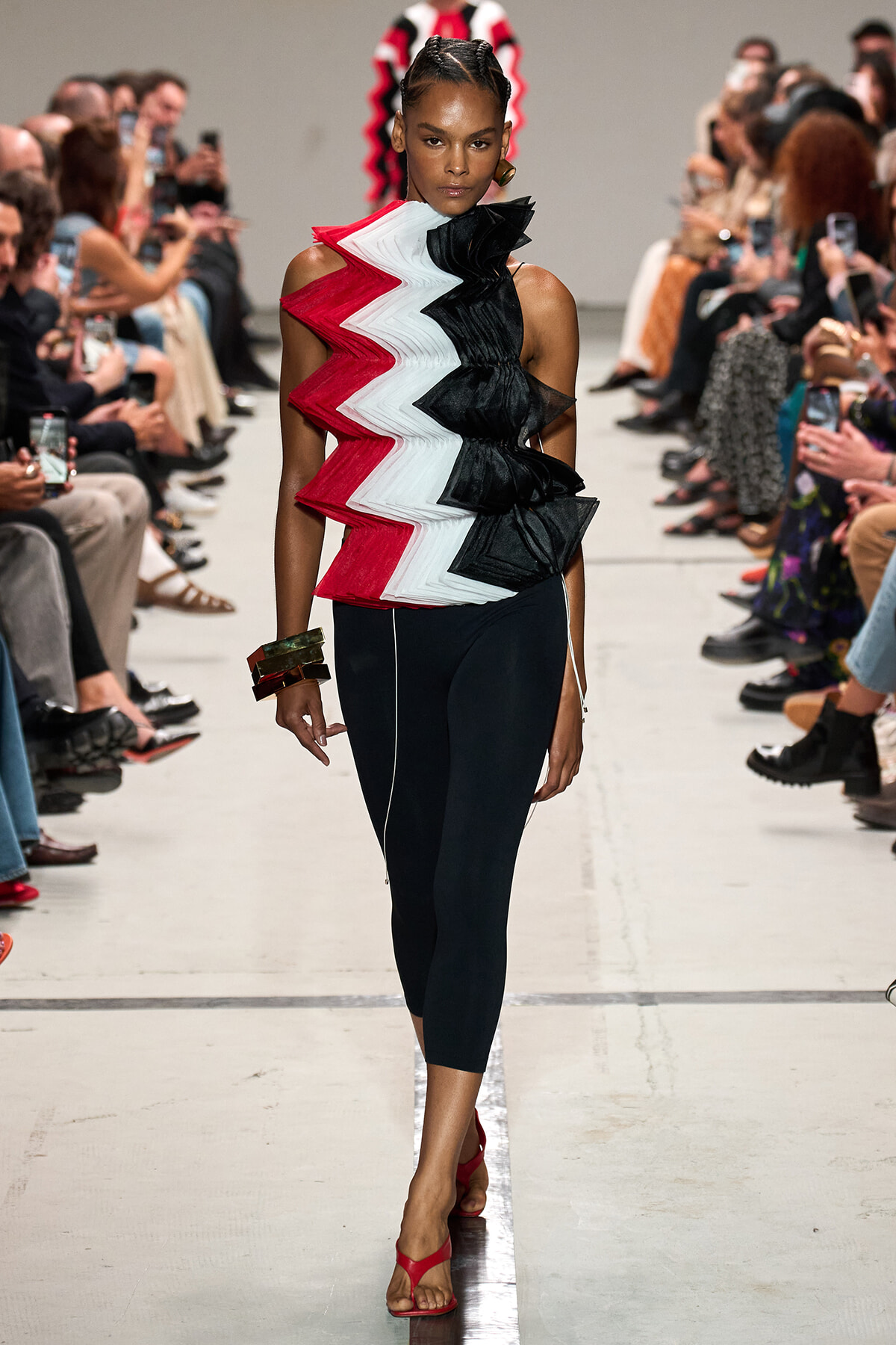 Model walking a fashion runway in a red, white, and black zigzag-pleated top with black leggings and red sandals, audience on both sides applauding/photo-taking.
