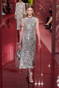 Model walking a runway in a full-length silver sequin dress covered with round mirrorlike discs, paired with metallic heels and a silver clutch.