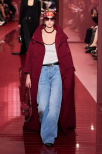 Model walking the runway in a burgundy patterned long coat over a white tank and light-wash wide-leg jeans, with a floral headscarf, sunglasses, and a burgundy handbag.