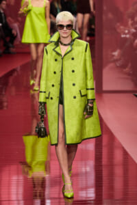 Runway model in a neon lime-green coat with black trim and a large collar, wearing sunglasses and gloves, lime heels, and carrying a small black handbag.