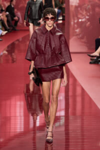 Model walks the glossy red runway in a metallic burgundy cocoon jacket and matching mini skirt, wearing oversized sunglasses and a clutch.
