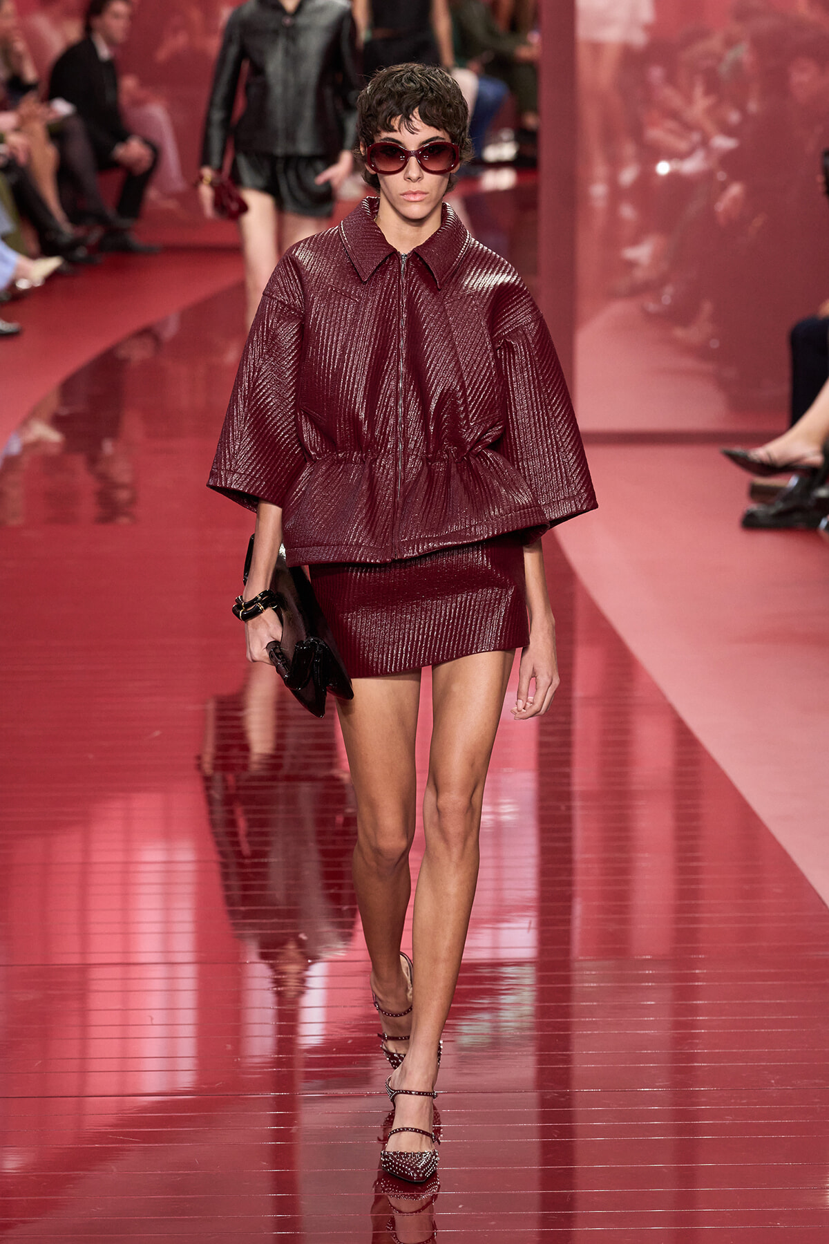 Model walks the glossy red runway in a metallic burgundy cocoon jacket and matching mini skirt, wearing oversized sunglasses and a clutch.