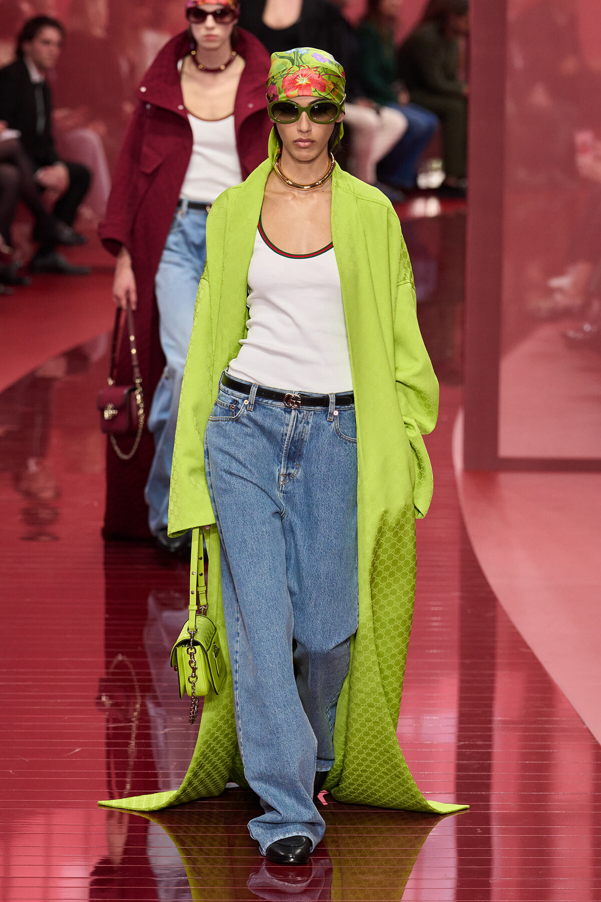 Model on a runway wearing a neon green coat over a white tank and loose jeans, with a colorful headscarf and sunglasses.