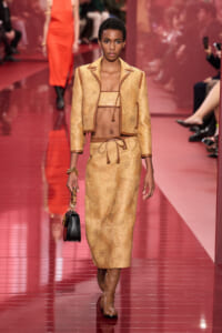 Model walking the runway in a tan lace two-piece set with a cropped top, matching jacket, and pencil skirt, carrying a small black handbag.