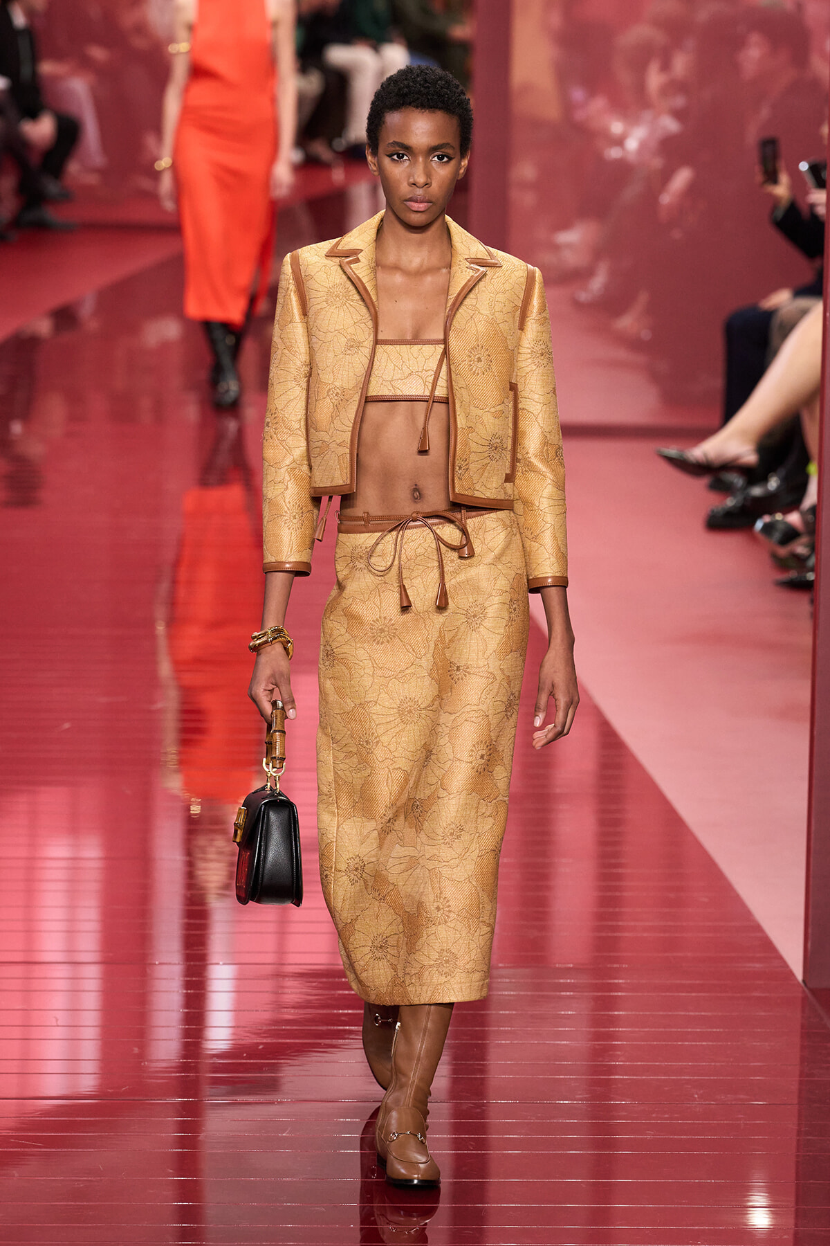 Model walking the runway in a tan lace two-piece set with a cropped top, matching jacket, and pencil skirt, carrying a small black handbag.