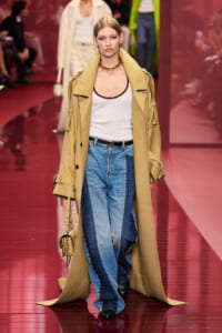 Model walks the pink runway wearing a long tan trench coat over a white tank with a green/red neckline, paired with baggy blue jeans and a beige handbag.