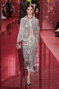 Model walks the runway in a silver sequined two-piece suit with a matching clutch and metallic heels on a pink catwalk.