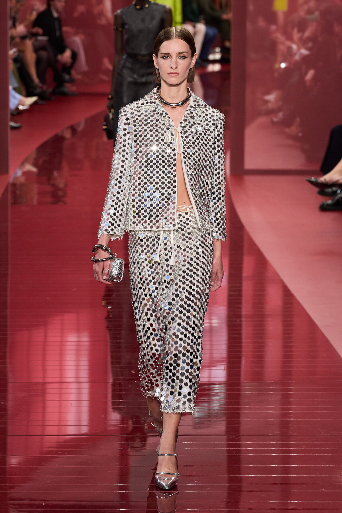 Model walks the runway in a silver sequined two-piece suit with a matching clutch and metallic heels on a pink catwalk.