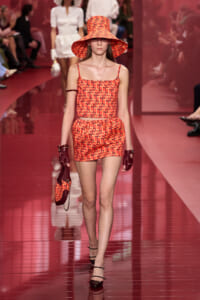 Model walks the runway in an orange geometric-print two-piece set with a wide-brim hat, burgundy gloves, and a matching handbag on a glossy red catwalk.