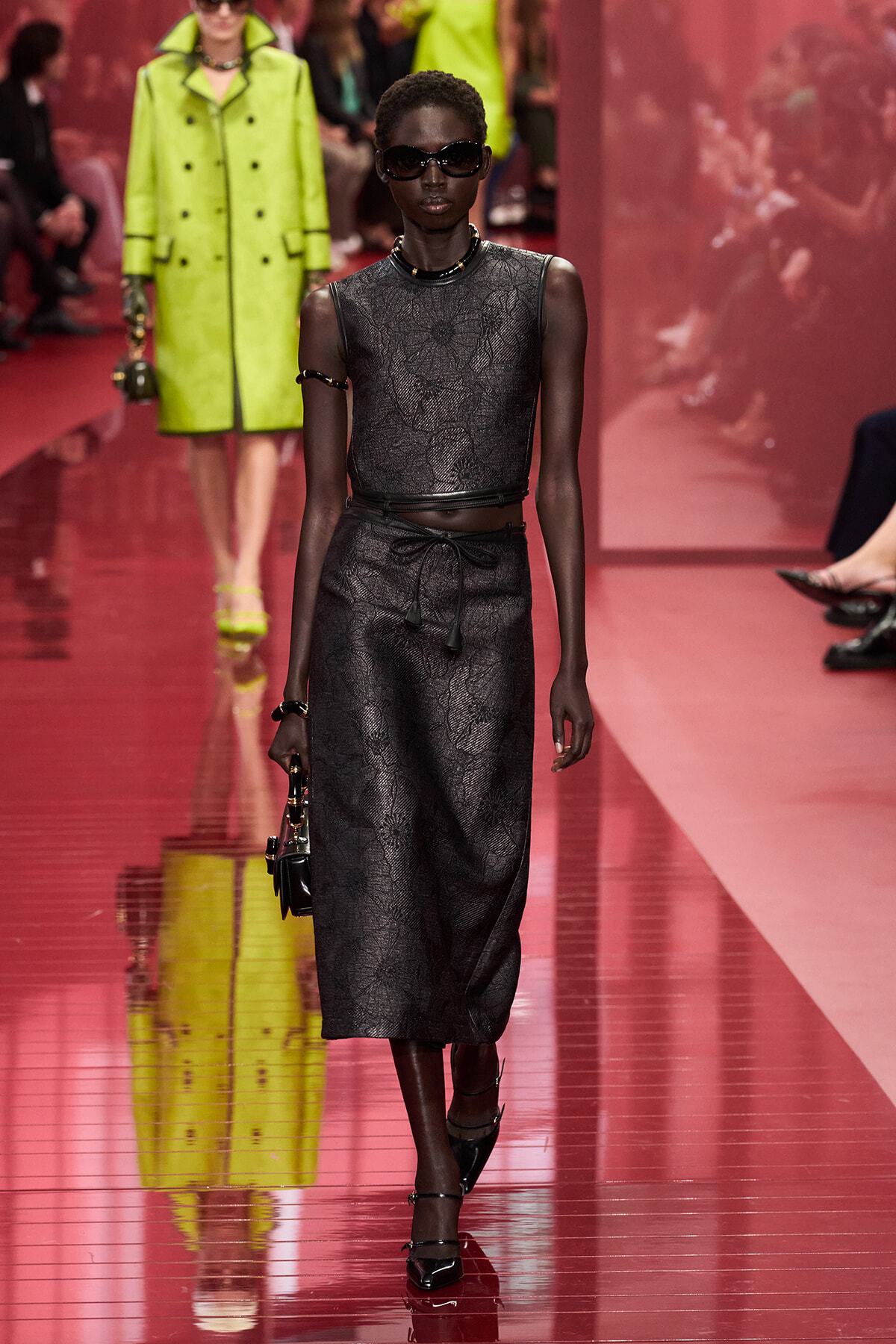 Model walks a pink runway in a black sleeveless lace-pattern two-piece set (crop top and midi skirt) with a belt, dark sunglasses, and a small black handbag. Neon green coat and audience blur in background.