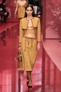 Model walking a fashion runway in a mustard yellow cropped jacket and knit set with brown belt, boots, and a small handbag.