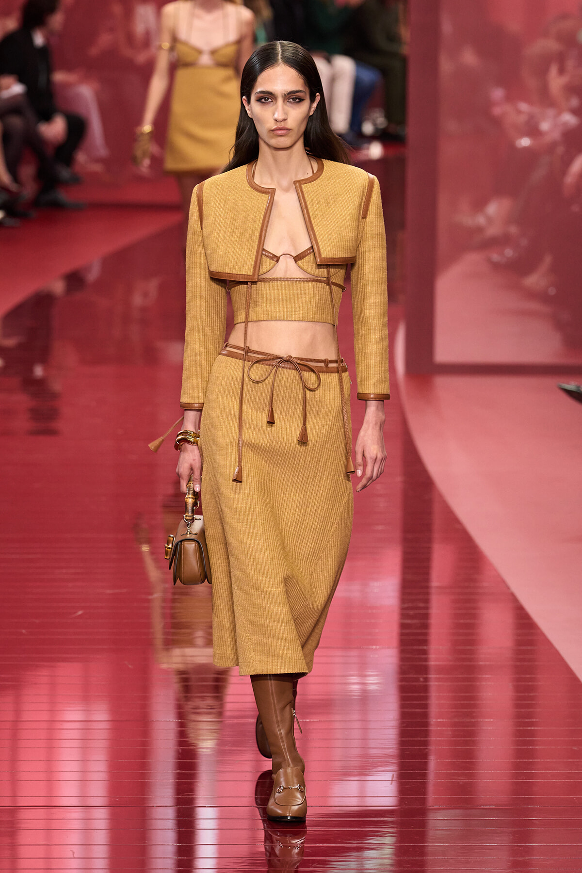 Model walking a fashion runway in a mustard yellow cropped jacket and knit set with brown belt, boots, and a small handbag.
