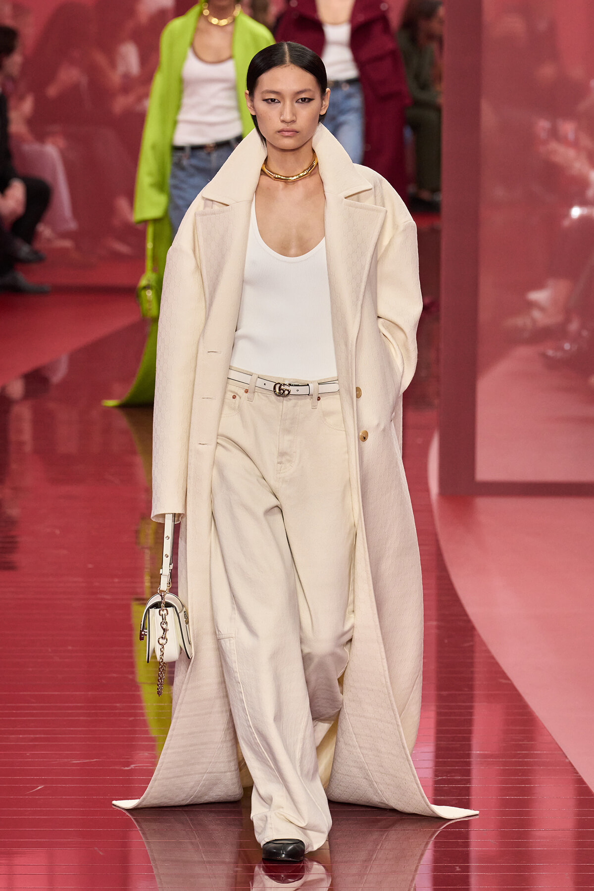 Model walks the runway in an oversized cream coat over a white tank, cream wide-leg pants with a beige belt, gold necklace, and a small white handbag.