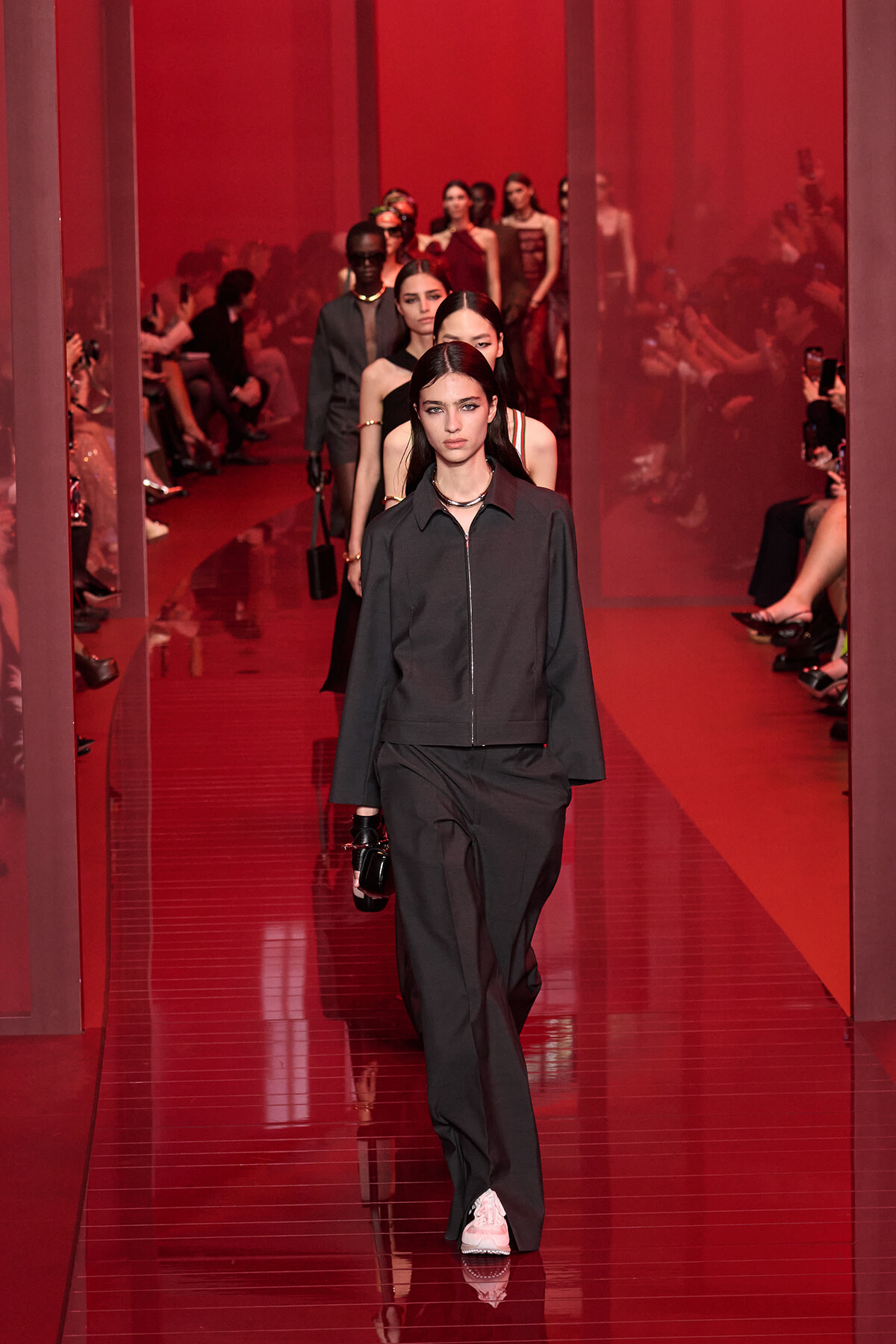 Model in a black zip-front jacket and wide-leg pants walks a glossy red runway, with other models following in line behind.