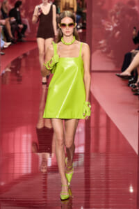 Model walks a neon lime patent-leather mini dress on a glossy pink runway, paired with matching gloves, choker, and sunglasses.