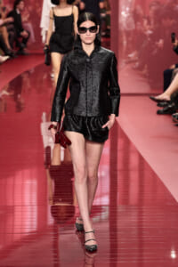 Model on a pink runway wearing a black textured jacket and matching shorts, with oversized sunglasses and a red clutch in hand.