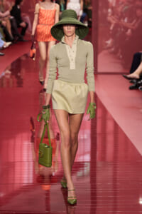 Model walks the runway in a beige long-sleeve top and matching mini skirt, with a wide green hat, green gloves, and a green shoulder bag on a pink glossy runway.