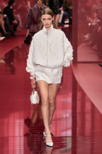 Model walks a runway wearing an oversized white textured jacket and matching white shorts, carrying a small white handbag and wearing white heels on a glossy pink runway.