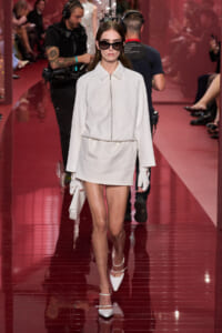 Model in a white zip-front coat and mini skirt, oversized sunglasses, white gloves, and pointed heels walking a glossy red runway.