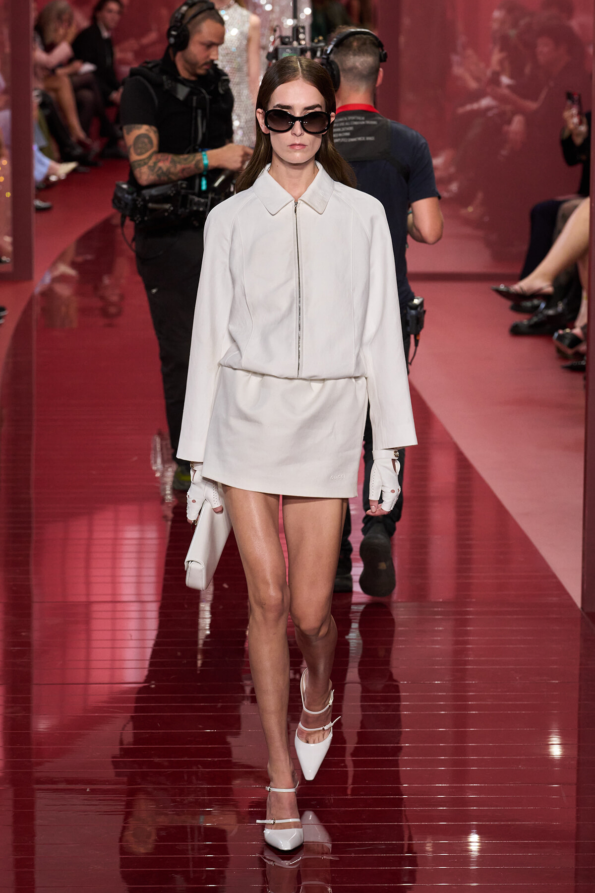 Model in a white zip-front coat and mini skirt, oversized sunglasses, white gloves, and pointed heels walking a glossy red runway.
