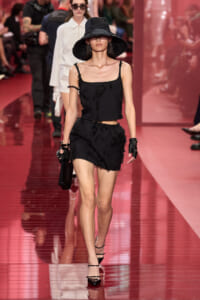 Model walking a fashion runway in a black textured crop top and frayed shorts, wide-brim hat, gloves, and a small black handbag.