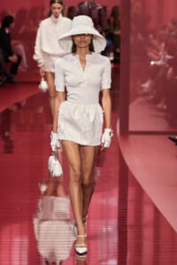 Model in white outfit with a wide-brim hat, short-sleeve top and ruffled mini skirt, white gloves, and white heels walks a pink runway while carrying a small white handbag.