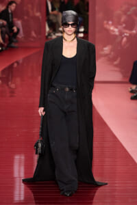 Model on a red runway wearing a long black coat over a black top and wide-leg jeans, with a patterned cap, dark sunglasses, a gold necklace, and a small black handbag.