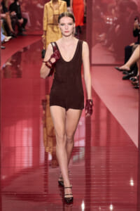Female model walking a fashion show in a sleeveless dark brown romper, red gloves, and a red handbag slung over her shoulder.