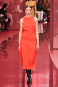 Model walking a fashion runway in a bright orange sleeveless midi dress with gold hardware, paired with black knee-high boots.
