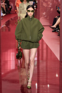 Model walks the runway in an oversized olive green zip-front jacket with matching shorts, green sunglasses, and a small dark green handbag.