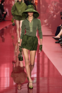 Model walks the runway in olive green outfit with a wide-brim hat, matching gloves, and a small handbag under pink lighting.