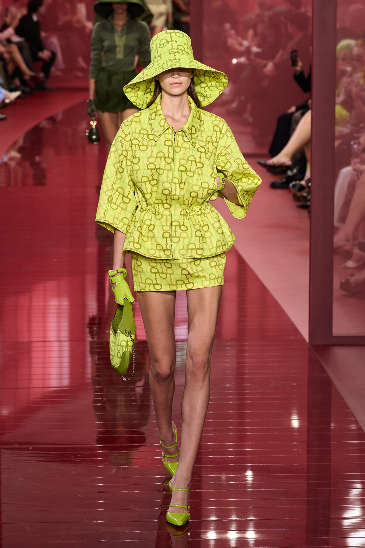 Model walking a fashion runway in a neon lime-green coordinated hat, jacket, and mini skirt with a matching handbag and gloves.
