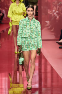 Model on a pink runway wearing a mint-green chain-pattern shirt and shorts set with a matching handbag and gloves.