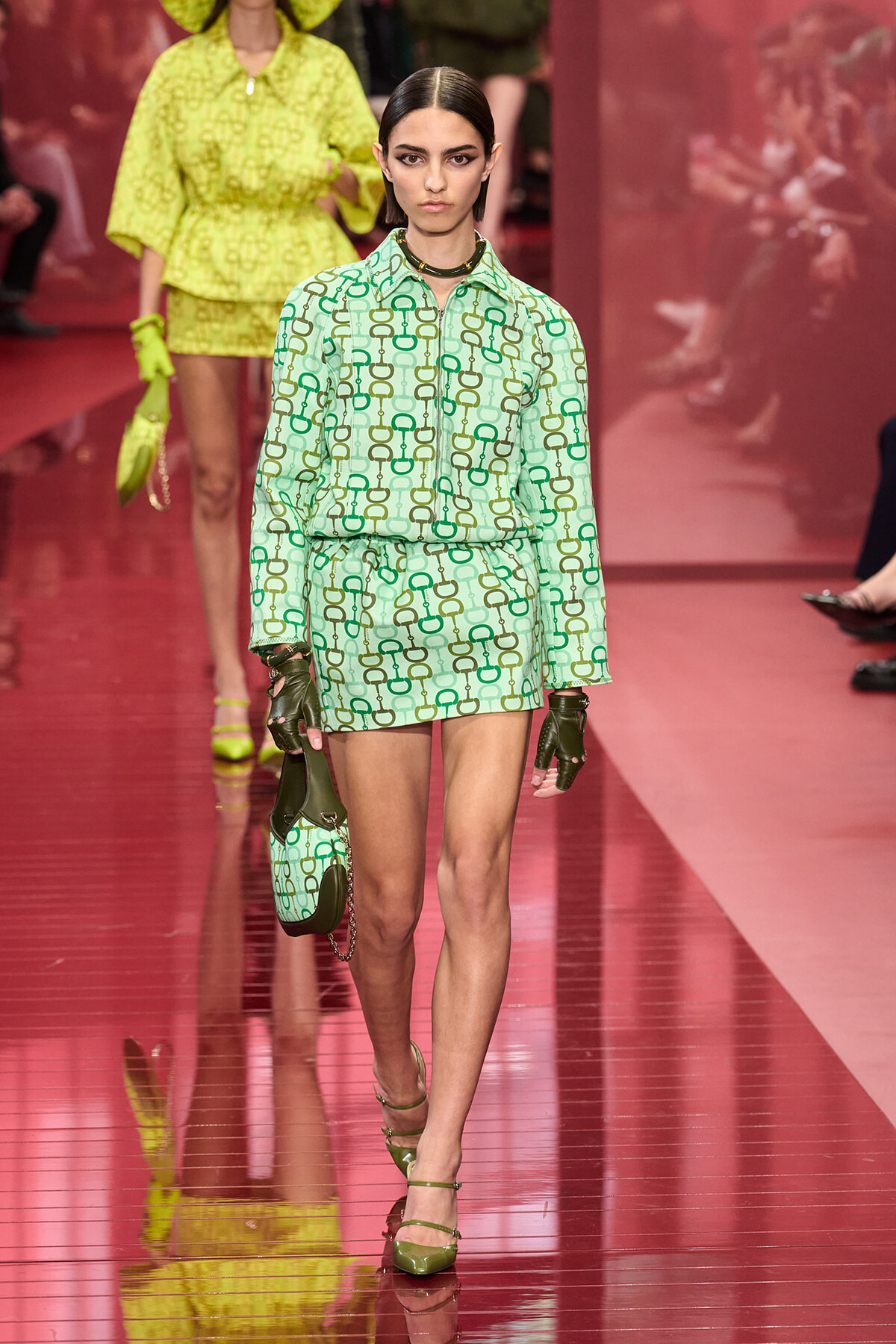 Model on a pink runway wearing a mint-green chain-pattern shirt and shorts set with a matching handbag and gloves.