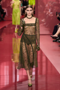 Runway model in a sheer black lace dress with floral patterns, olive-green gloves, and a green choker walking a pink catwalk.